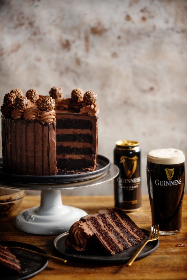 巧克力吉尼斯蛋糕放在蛋糕架上，切一片，放在盘子里。后面是一个空的吉尼斯黑啤酒罐和一个满杯子的吉尼斯黑啤酒。